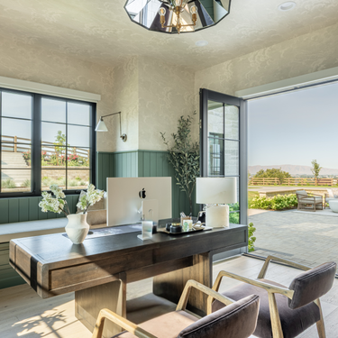 Modern home office with a desk, computer, and chairs, featuring large windows with a view of greenery.